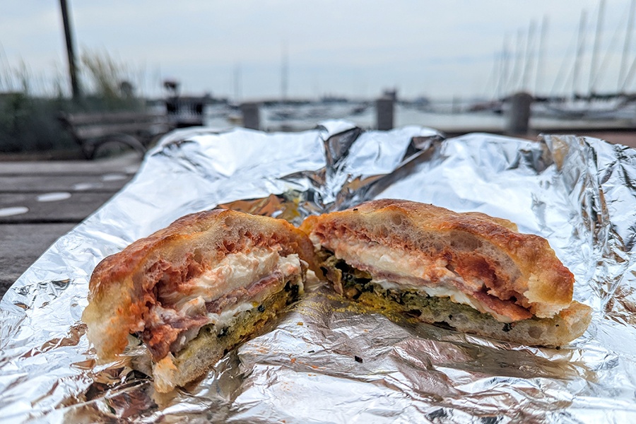 A breakfast sandwich with eggs, pesto, tomato jam, and more on focaccia is photographed outdoors with water and boats in the background.