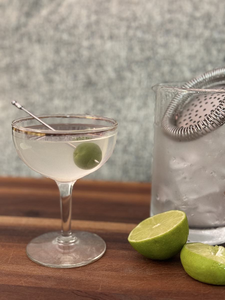 A clear cocktail is garnished with a green olive and photographed next to sliced lime.