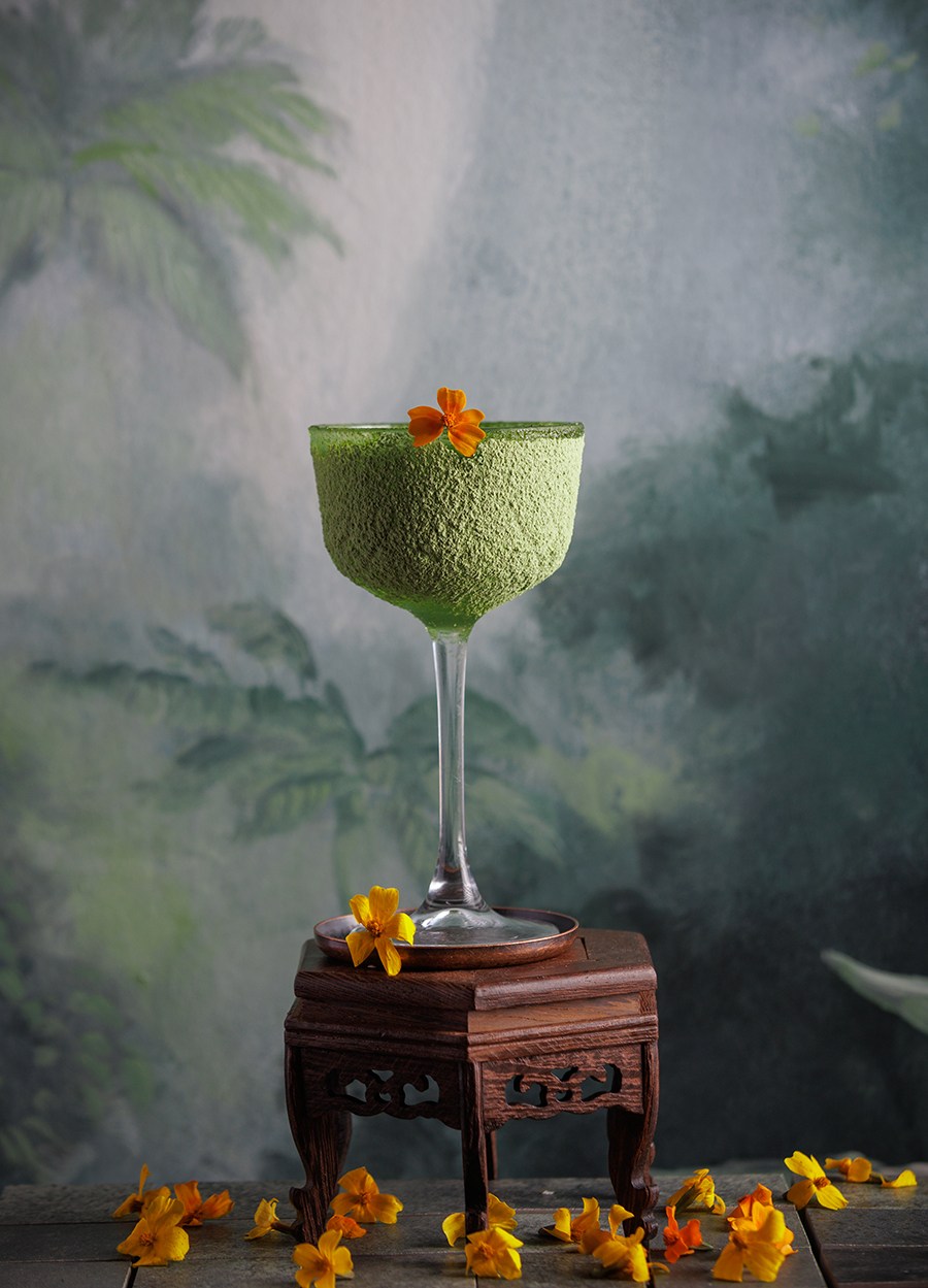 A green cocktail is displayed in front of a wall painted with palm trees and garnished with edible flowers.