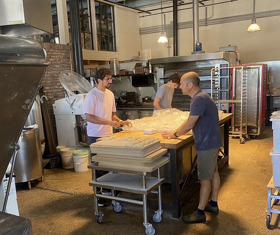 Three people work in a spacious bakery, shaping bagels.