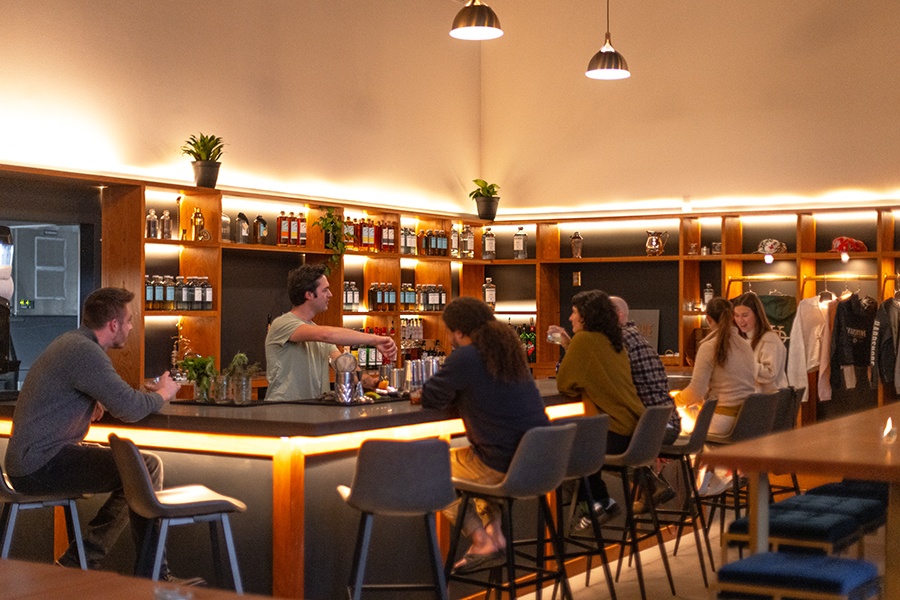 People sit at a bar inside a distillery with lots of light wooden surfaces and some plants.