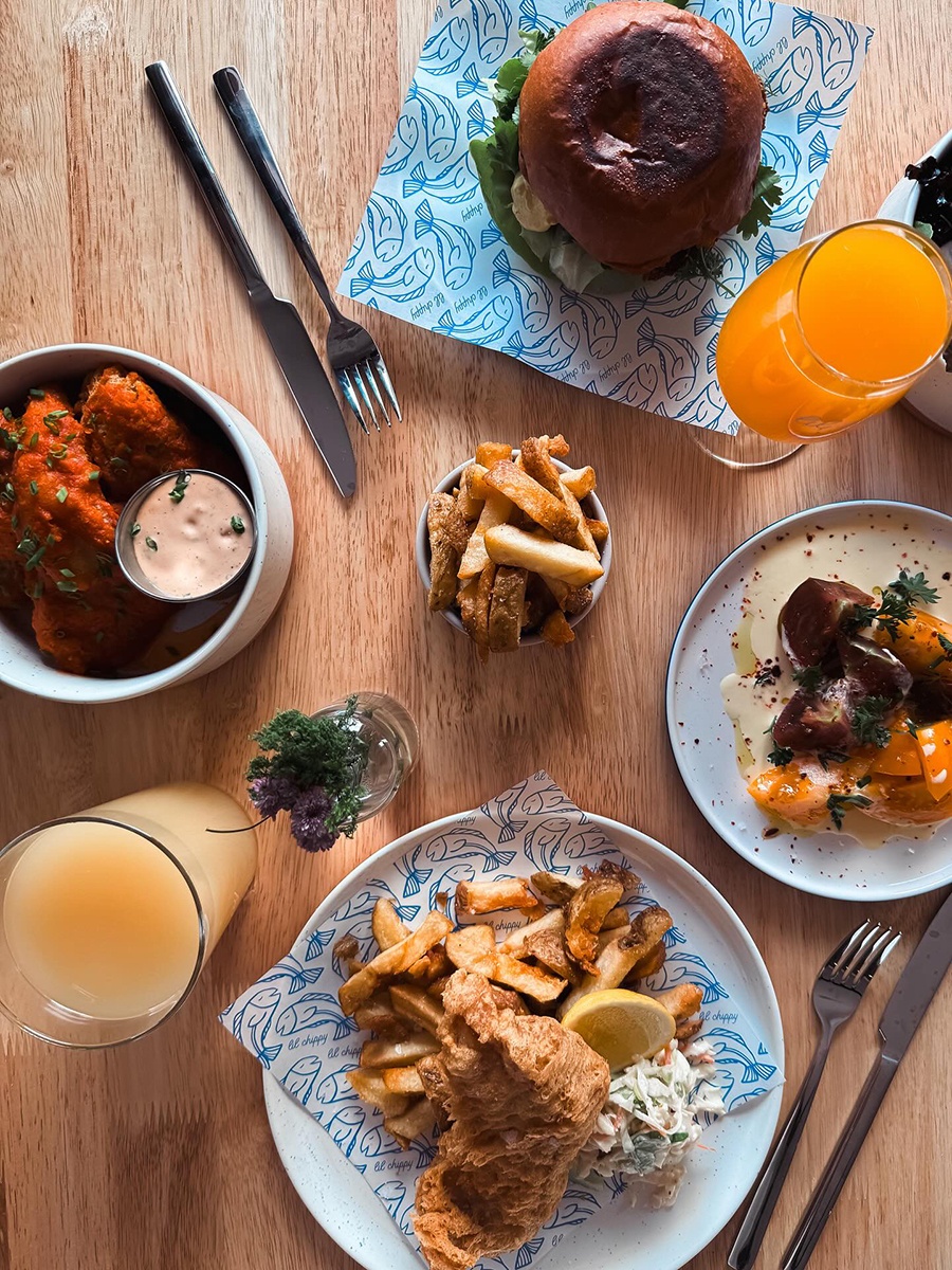 Overhead view of fish and chips and other food on a light wooden table.