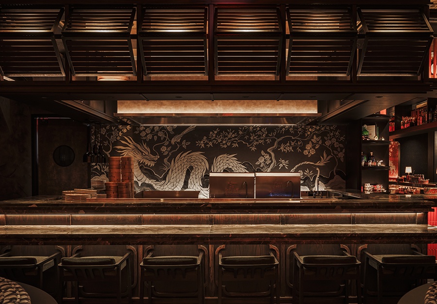 A dragon mural in brown and beige tones sits behind a restaurant bar area with lots of dark wood.