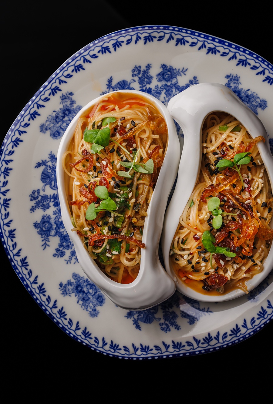 Oysters and vermicelli topped with microgreens and sesame seeds are served in two oyster-shaped bowls atop a white plate with a blue floral design.