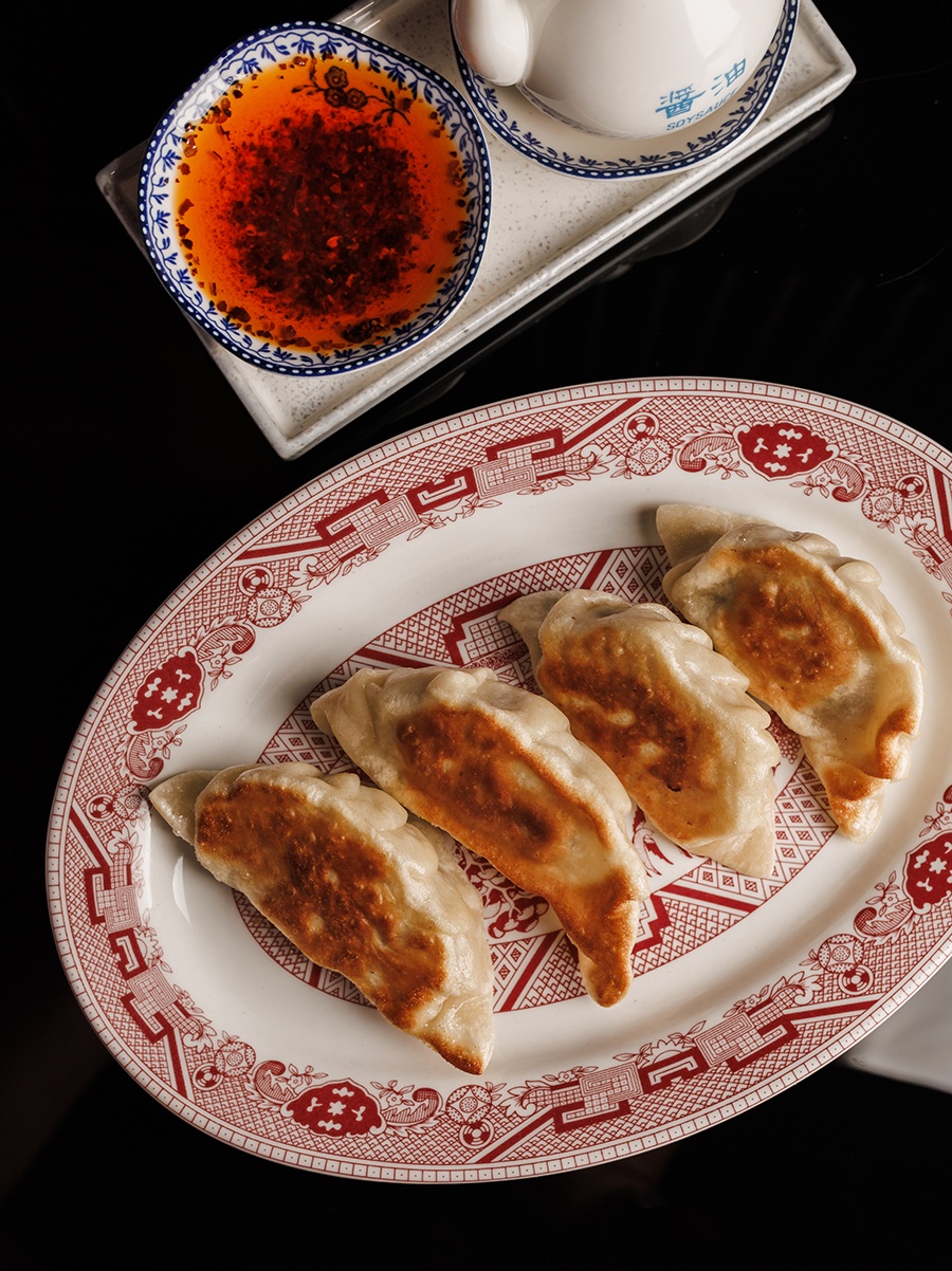 Pan-seared dumplings sit on a red and white plate, accompanied by a little dish of a chili oil.