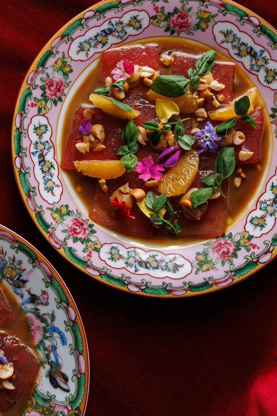 Raw tuna is garnished with herbs, edible flowers, and tangerine slices on a colorful floral plate.