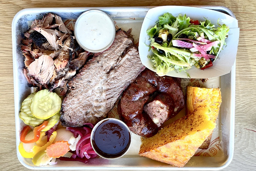 Overhead view of a barbecue platter that includes brisket, sausage, cornbread, pickles, and more.