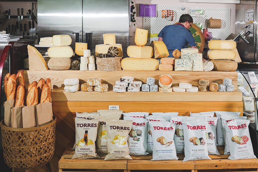 Cheese-filled interior of a cheese shop, with baguettes and Torres potato chips also visible.