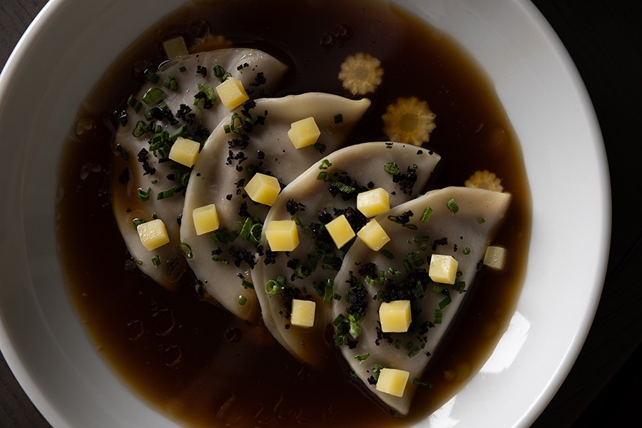 Four dumplings sit in a dark brown broth, topped with small cubes of cheese.