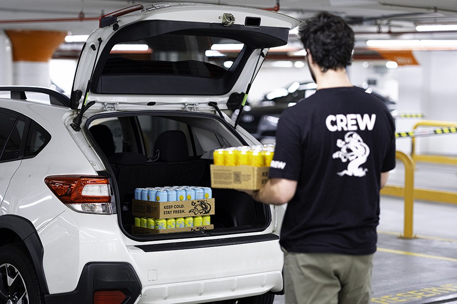 A person in a t-shirt that says "crew" on the back loads cans of beer into the open trunk of a car inside a parking garage.