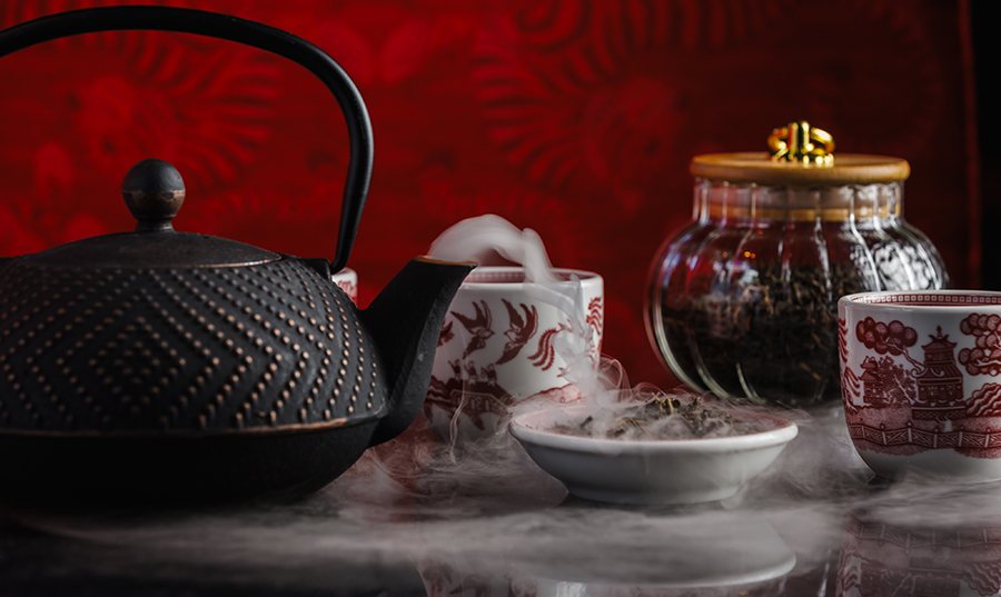 Steam pours out of a Chinese-style tea pot, displayed next to red and white tea cups.