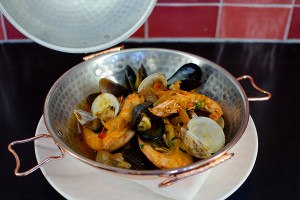 Clams, mussels, shrimp, and broth sit in a big copper pot in front of a red tiled wall.