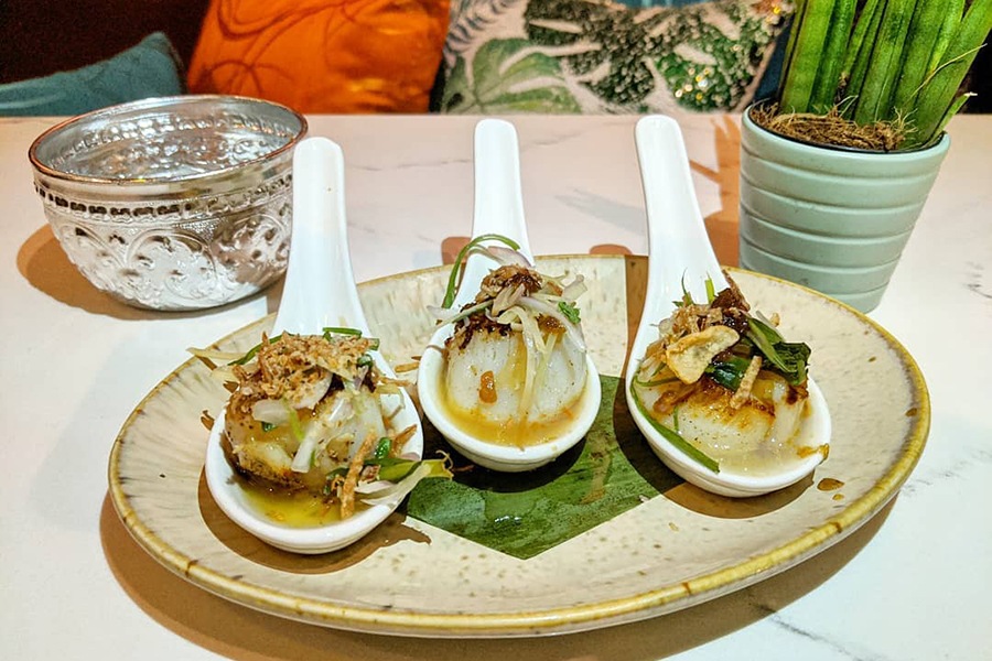 A seared scallop sits in each of three large white soup spoons.