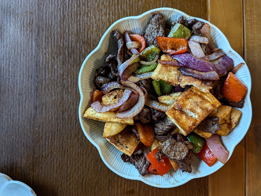 Overhead view of a stir-fried dish of lamb strips, chunks of flatbread, red onions, and bell peppers.