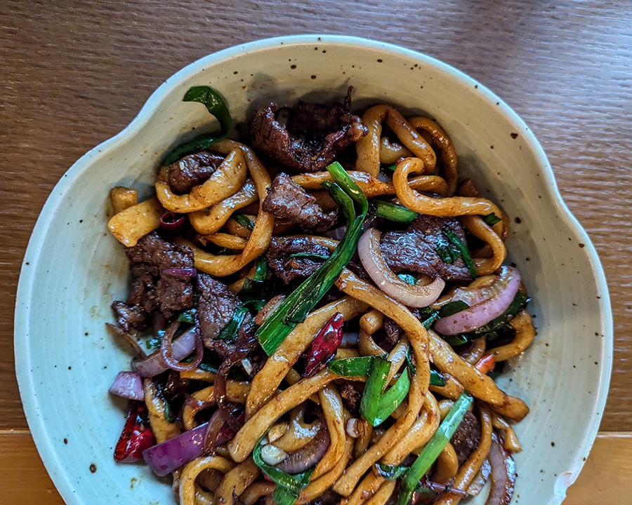 Overhead view of a bowl of medium-width noodles stir-fried with beef, red onions, and chives.