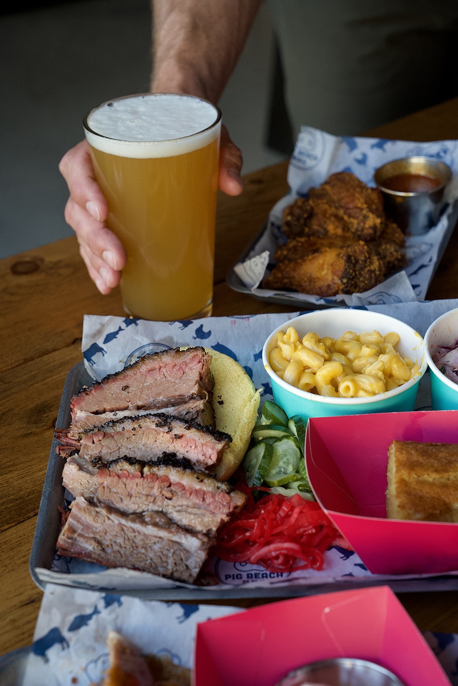 A hand holds a pint of beer on a table among brisket, mac and cheese, and other barbecue items.