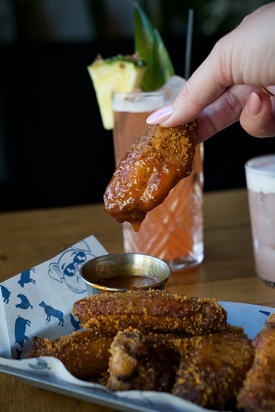 A hand dips a dry-rubbed chicken wing into an orange-brown sauce, with a pineapple-bedecked cocktail visible in the background.