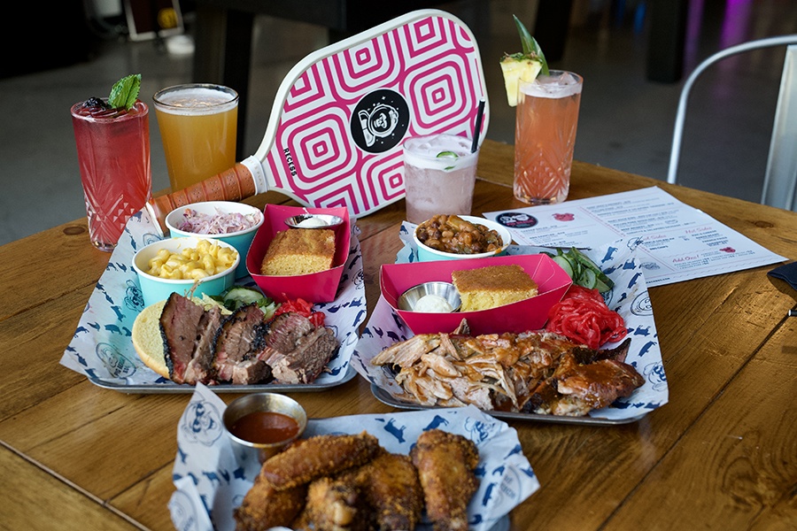 Trays of barbecue food sit on a wooden table with cocktails, beer, and a pickleball paddle in the background.