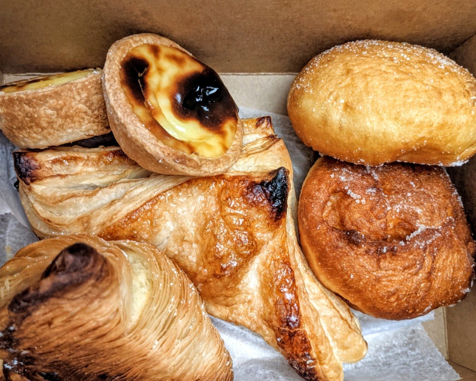 A variety of pastries, including egg tarts, sit on tissue paper in a cardboard takeout box.