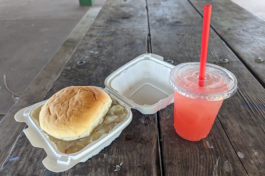 Saucy beansprouts between buns fill a white styrofoam container next to pink lemonade on a picnic table.