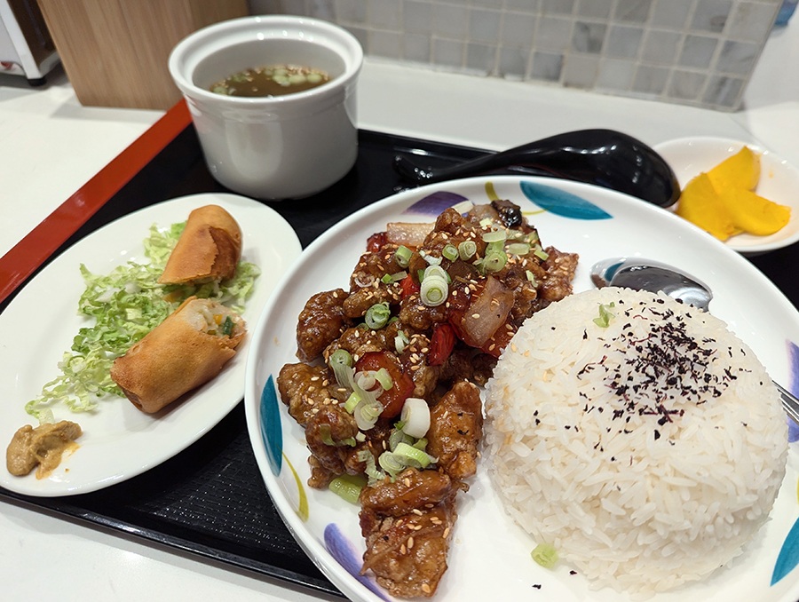 Crispy chicken, white rice, a veggie roll, and soup sit on a black tray.