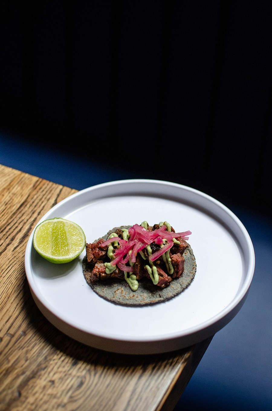 A blue corn tortilla is topped with chunks of beef, red onions, and a squiggle of avocado crema.