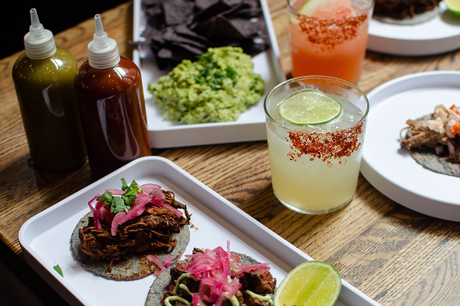 Tacos, colorful cocktails, and guac with blue corn chips sit on a wooden table.