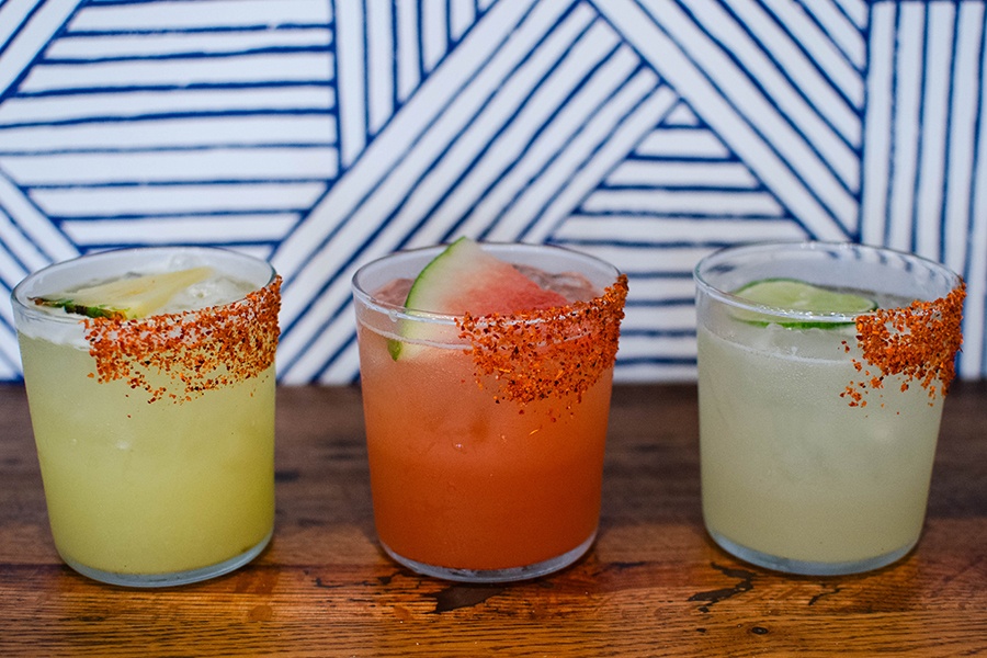 A trio of cocktails are lined up in front of a wall with a blue and white striped pattern.