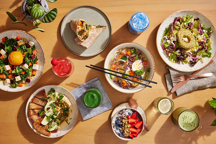 Overhead view of salad, soup, wrap, acai bowl, and smoothies on a light wooden table.