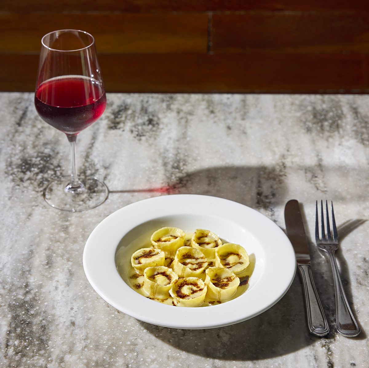 A marble tabletop is set with a white bowl of filled pasta drizzled with balsamic, accompanied by a glass of red wine.