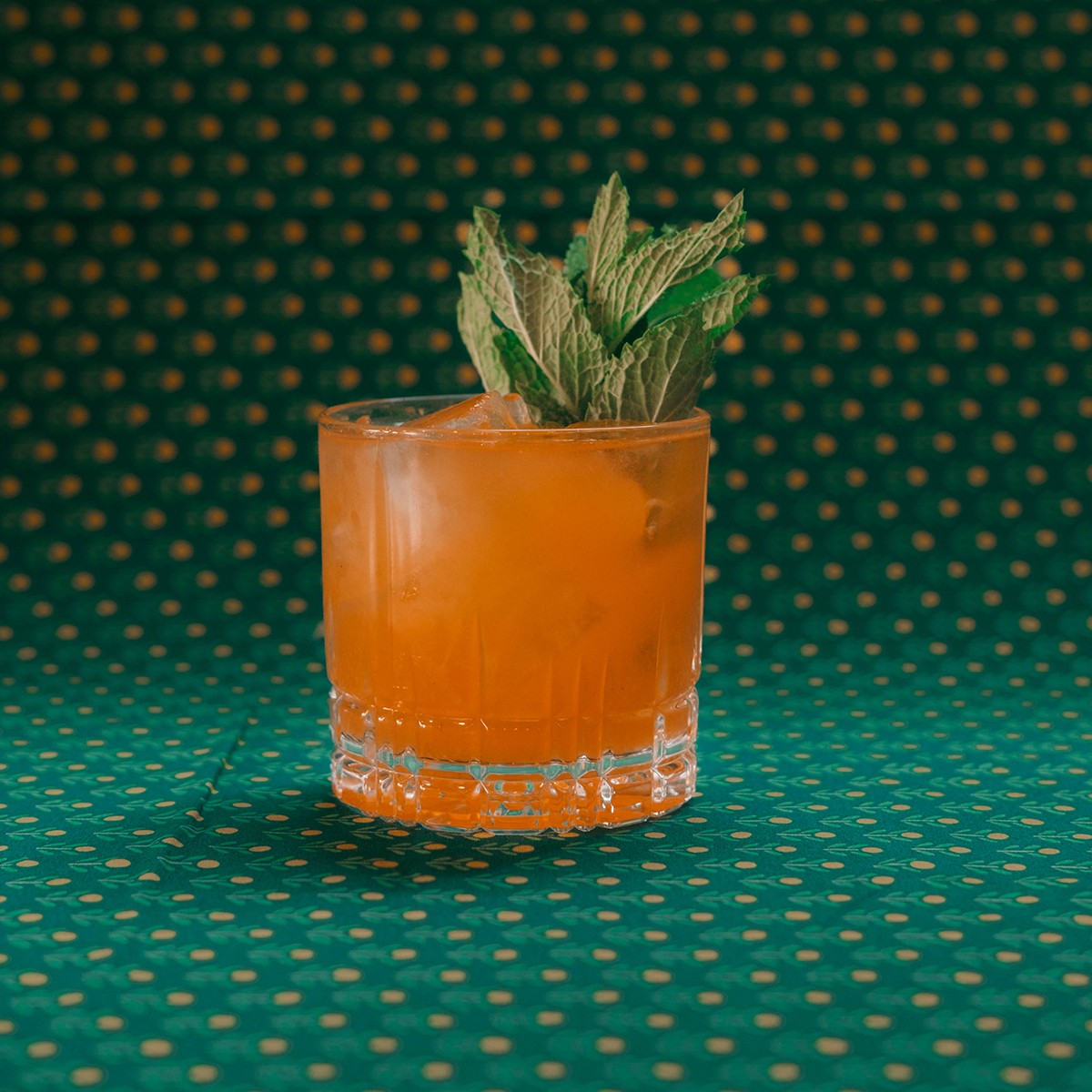 An orange cocktail sits on a patterned green background.