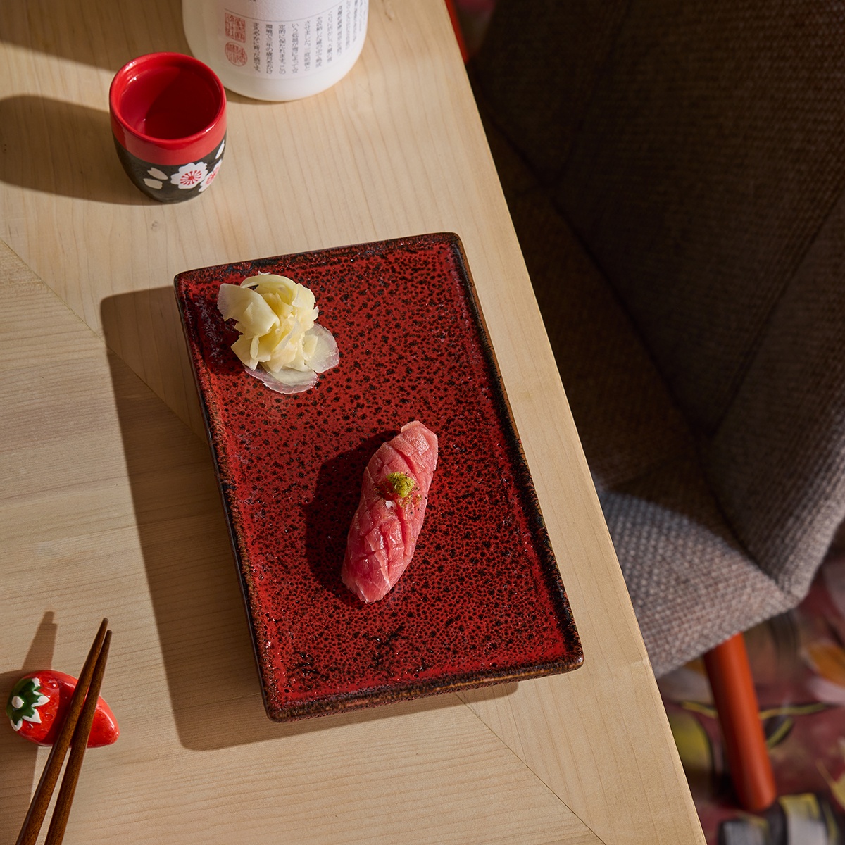 Overhead view of a single piece of nigiri on a red plate with chopsticks and a sake cup in view.