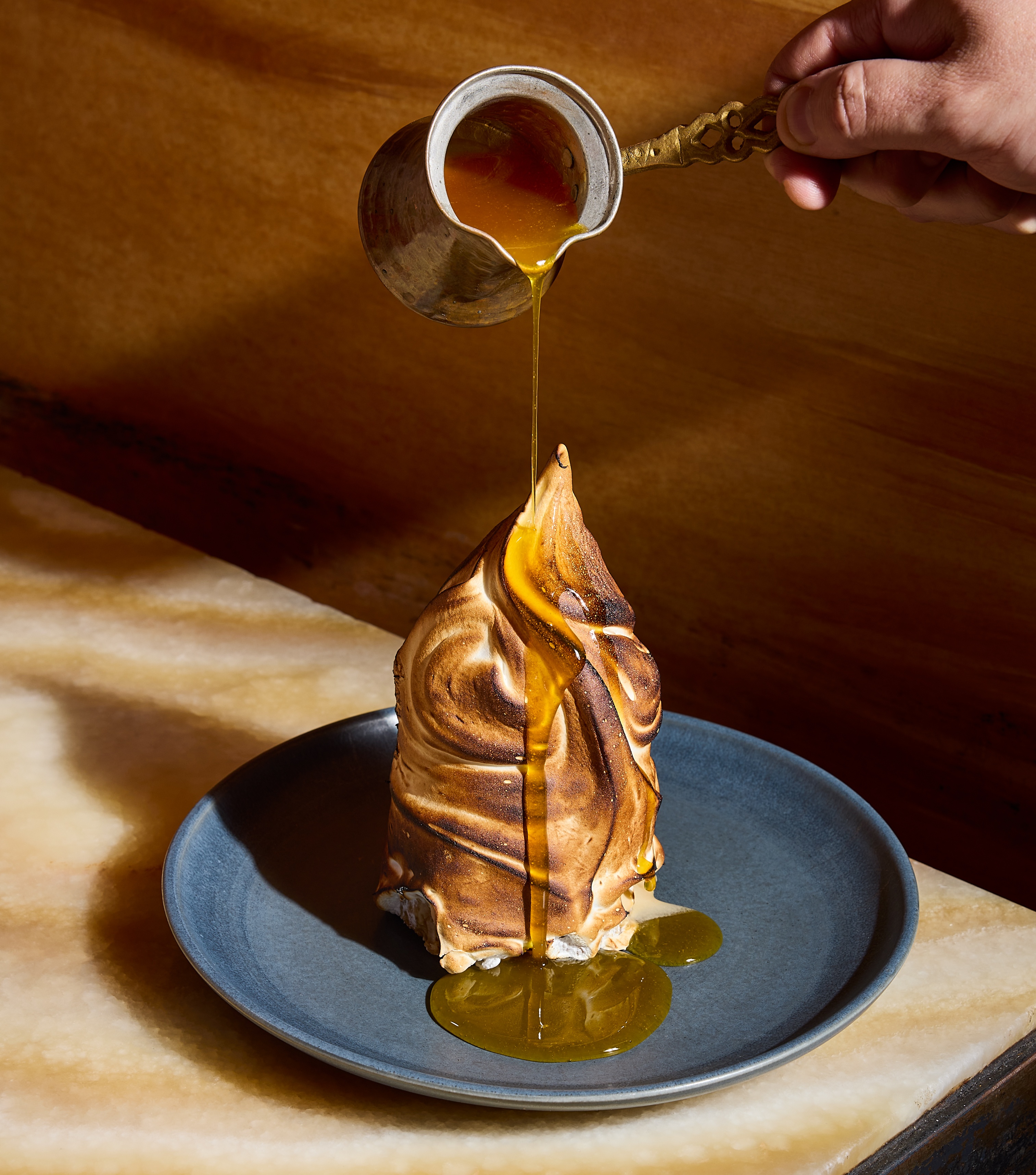 A drizzle of caramel is poured down over the toasted-meringue exterior of a baked Alaska dessert, which is sitting on a blue plate.