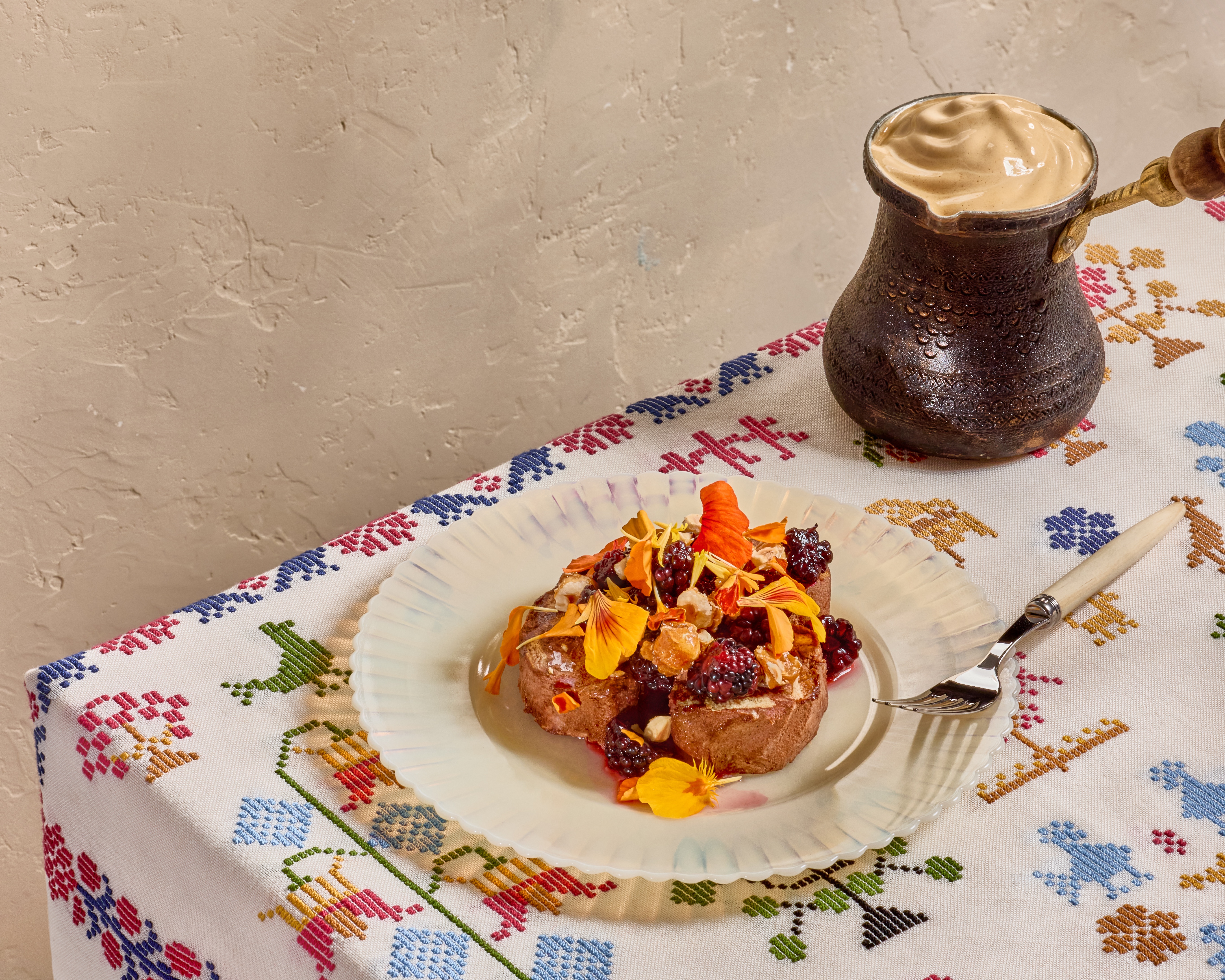 A dessert covered with edible flowers sits on a colorful tablecloth featuring Greek-style embroidery.