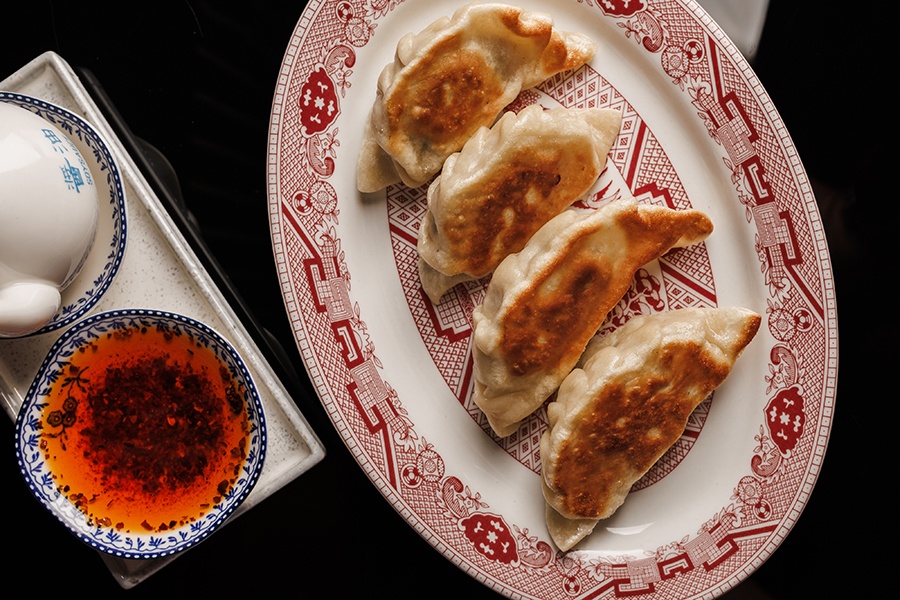 Pan-seared pork dumplings are lined up on a red and white plate next to a bowl of chili oil.