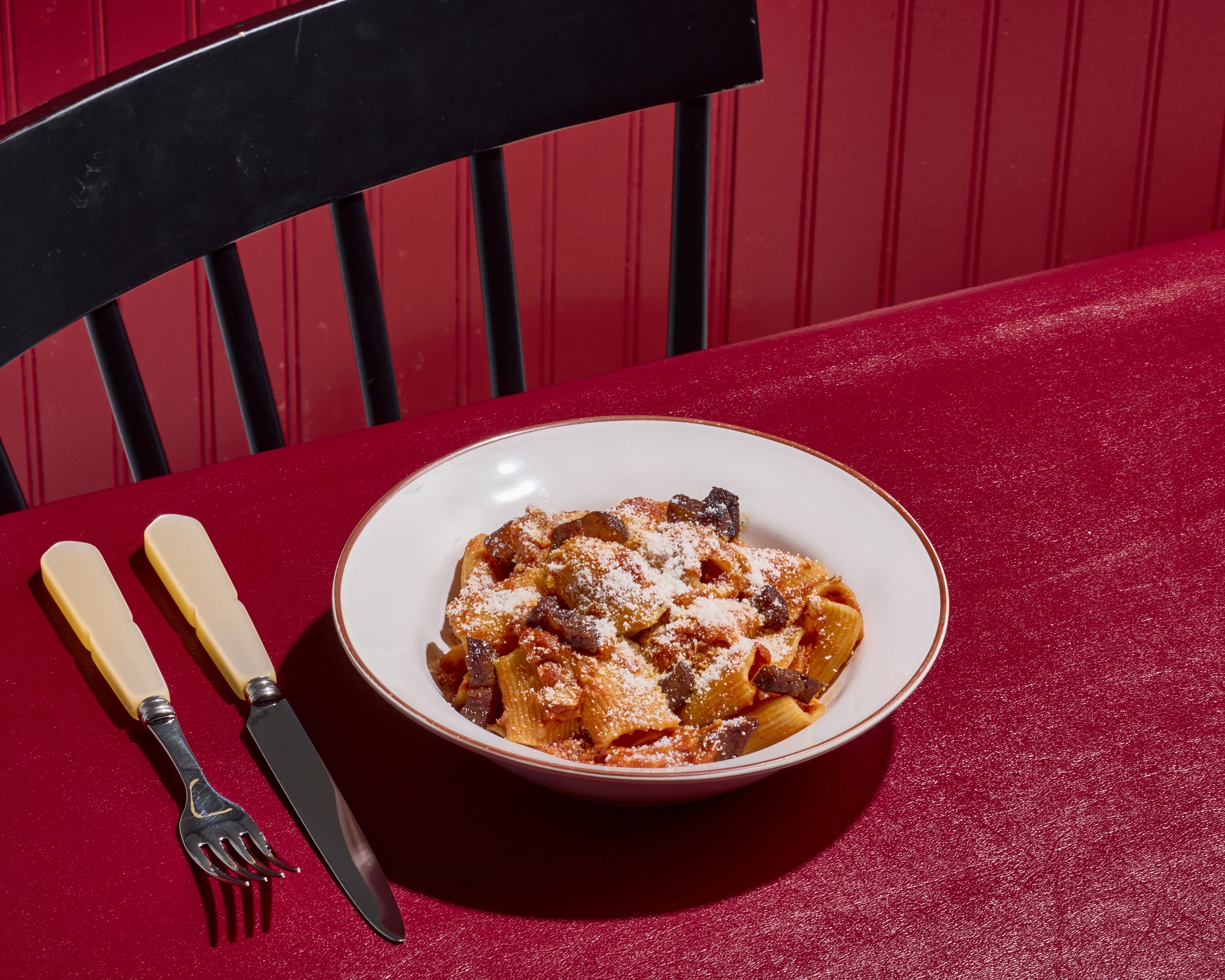 A white bowl of rigatoni Amatriciana on a red vinyl tabletop with a black chair and a read wall from Tonino in Jamaica Plain.