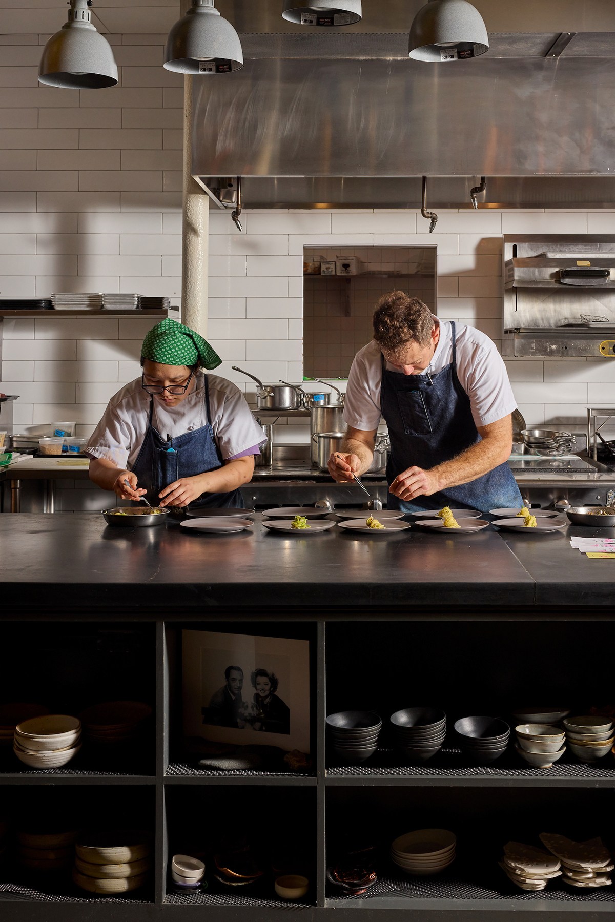 Two chefs in blue aprons concentrate while plating elegant dishes in an open kitchen.