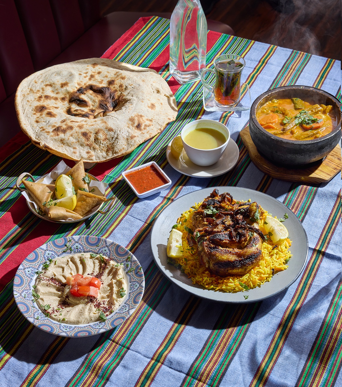 A colorful striped tablecloth is covered with a spread of Yemeni dishes, including grilled chicken over rice, triangular crispy samboosas, a steaming bowl of stew, and a giant round of flatbread.