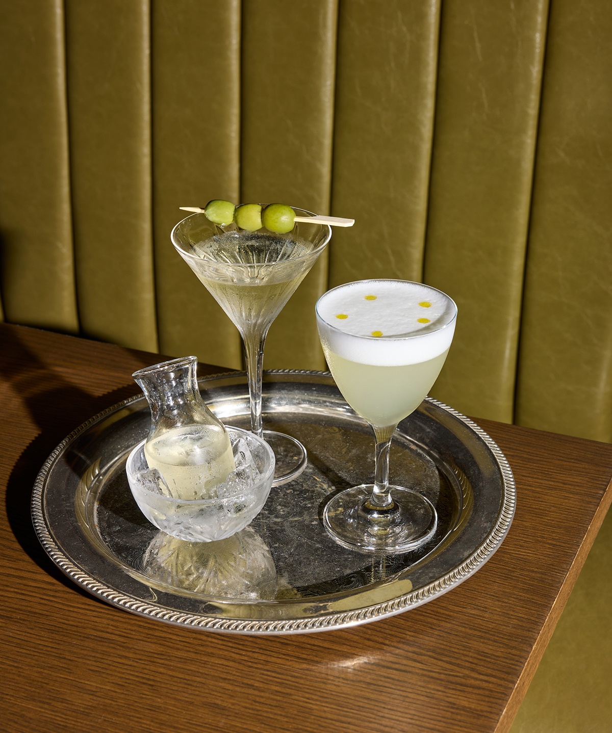 A martini and other cocktails are displayed on a silver tray on a restaurant table with an olive green booth.