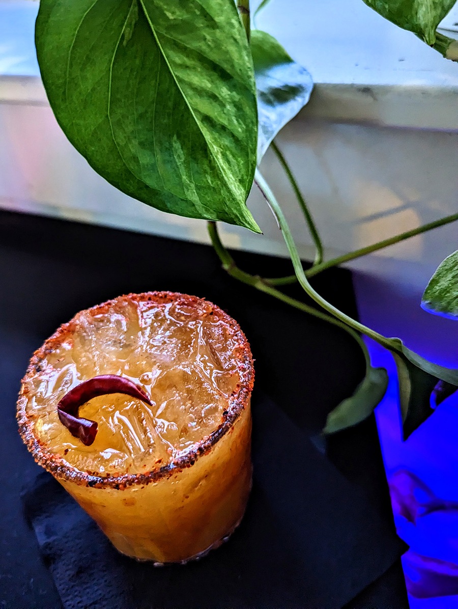 An orange cocktail with a chili pepper and chili salt rim sits on a black bar.