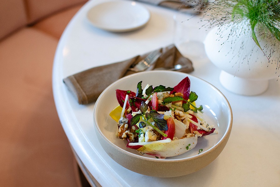 A salad of endives, cheese, and greens is served in a bowl on a white marble table.