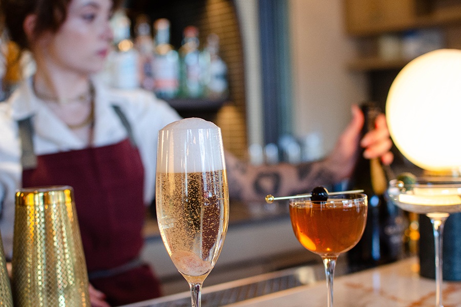 A bartender in a red apron stands behind a bar with several cocktails in front of her.