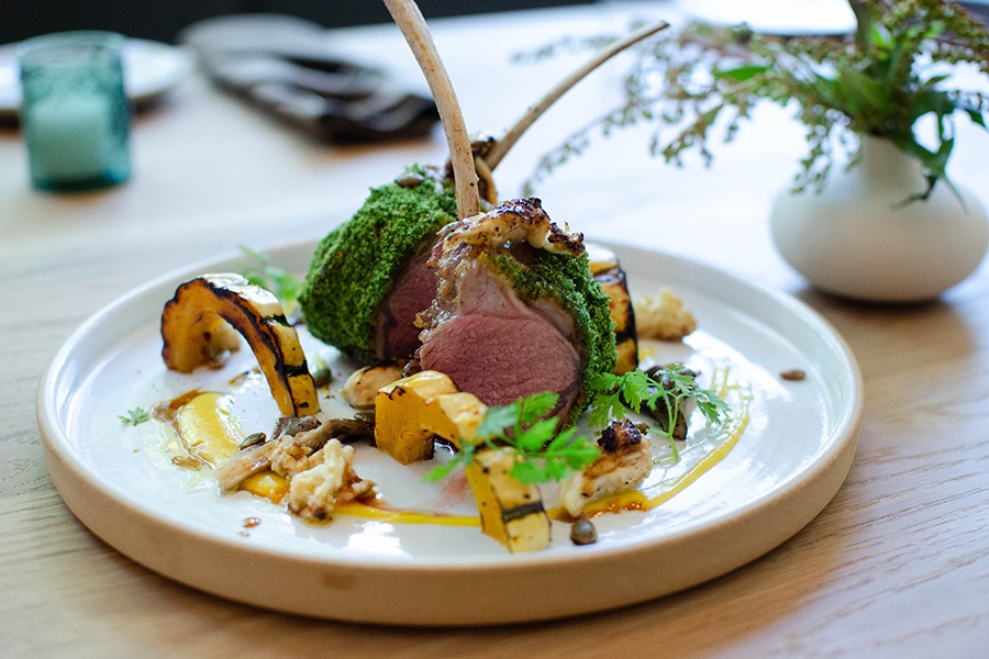 Lamb chops with a bright green herb crust are standing up tall on a plate with curves of squash and a swoosh of sauce.