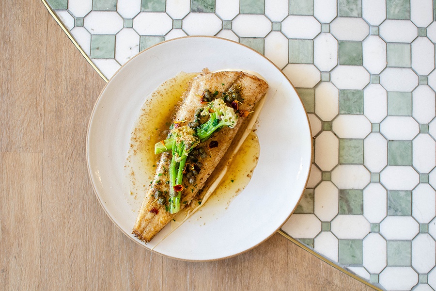 Overhead view of a sole filet in a brown butter sauce, served on a white plate, which is sitting on a tiled floor.