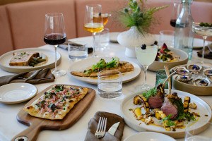 Lamb, oysters, tarte flambee, sole meuniere, and more dishes are spread across a white marble table, accompanied by wine and cocktails.