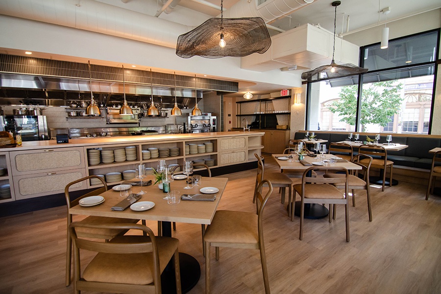 A restaurant, photographed empty during the day, features lots of light wood and a big open kitchen.