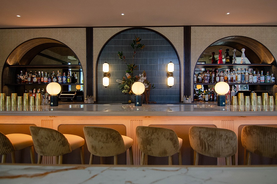 A restaurant bar features a white marble bartop, light brown velvet seats, and arched shelving.