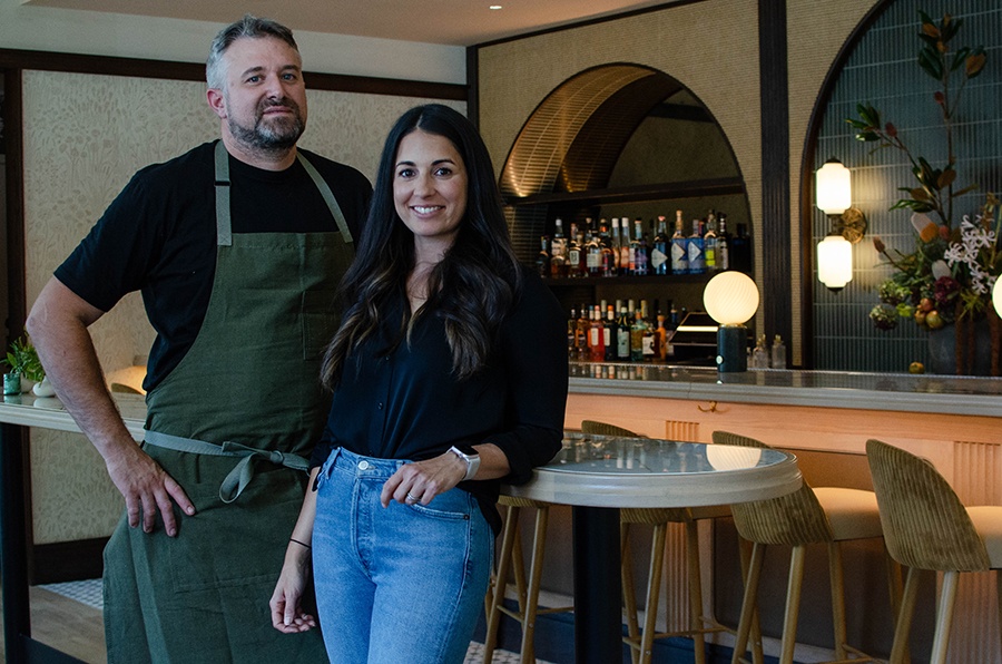 A man in a chef's apron and a woman in jeans stand smiling in a restaurant.