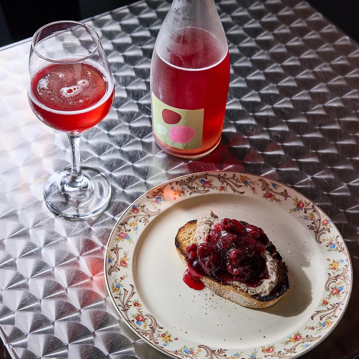 A piece of toast is topped with a seafood spread and sour cherry preserves, accompanied by a glass of bold pink wine.