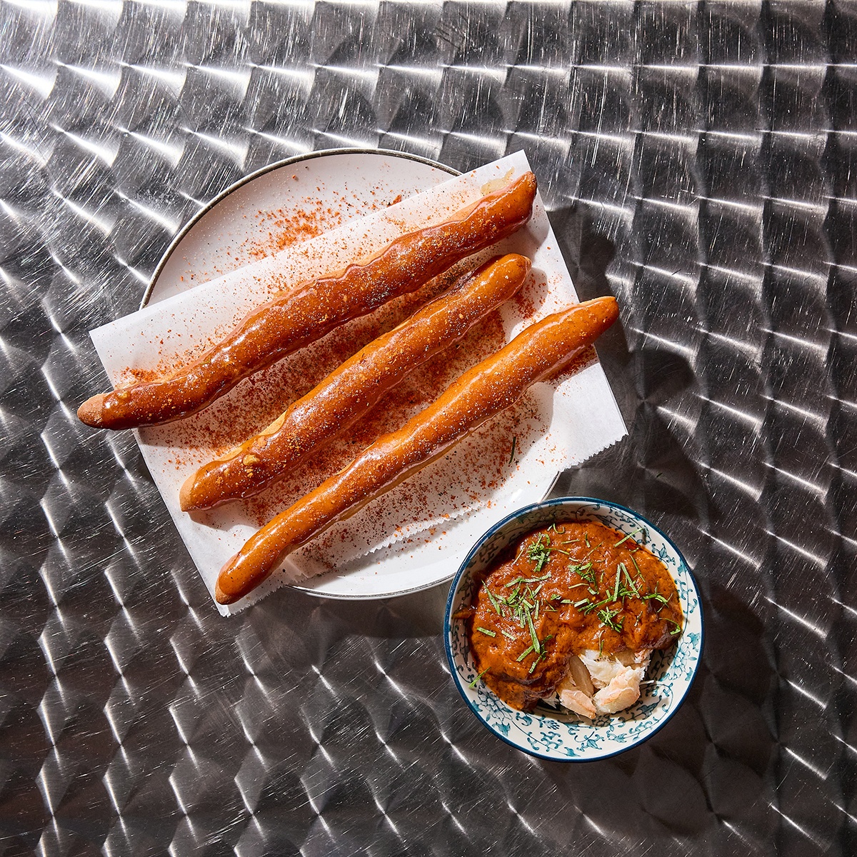 Overhead view of three long, thin pieces of fried dough with a side of crab in a brown curry sauce on a silver table.